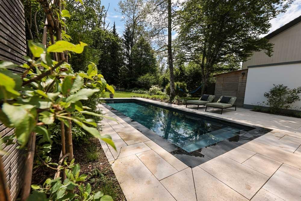 Piscine naturelle design avec bains de soleil et terrasse en pierre claire dans un jardin vert