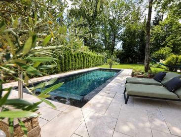 Piscine naturelle design avec terrasse en pierre claire et bains de soleil dans un jardin calme