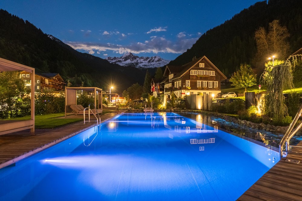 Piscine d’hôtel éclairée la nuit devant un village de montagne et les Alpes