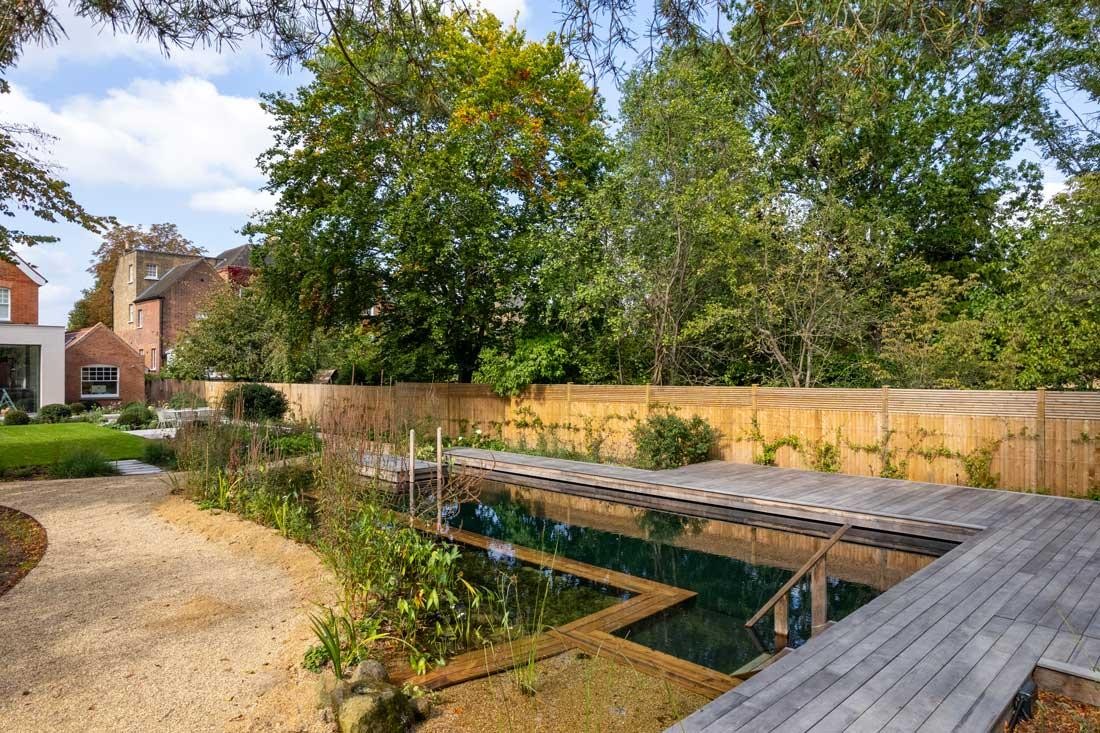 Piscine naturelle avec ponton en bois devant un pavillon de jardin moderne dans un jardin verdoyant
